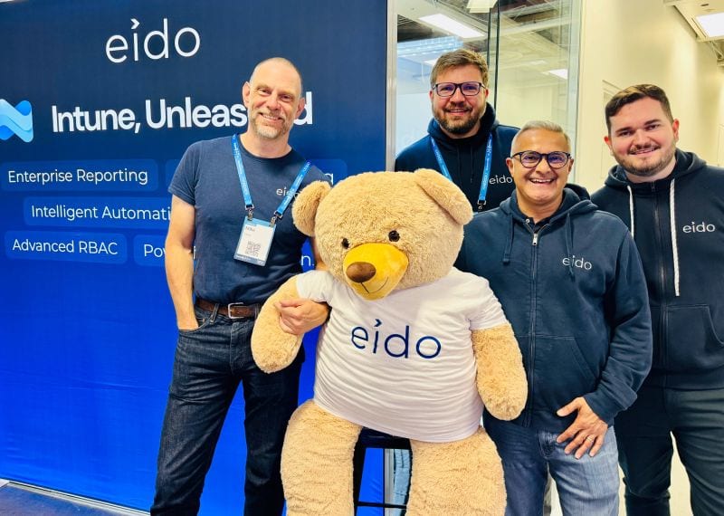 Four of the Eido team with Odie, a large teddy bear in an Eido T-shirt, in front of the Eido stand at Experts Live UK.