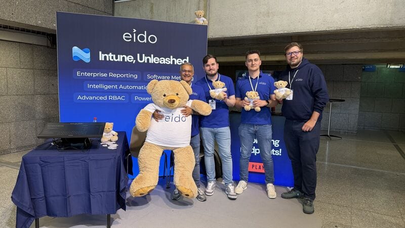 Four of the Eido team and Odie, a large teddy bear in an Eido T-shirt, in front of the Eido stand at MEM Summit.